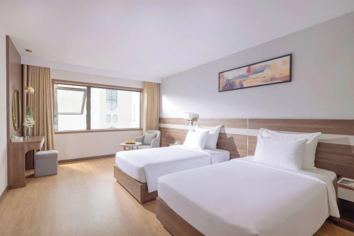 A hotel room with two beds, a desk, a chair, a window, and a painting on the wall.