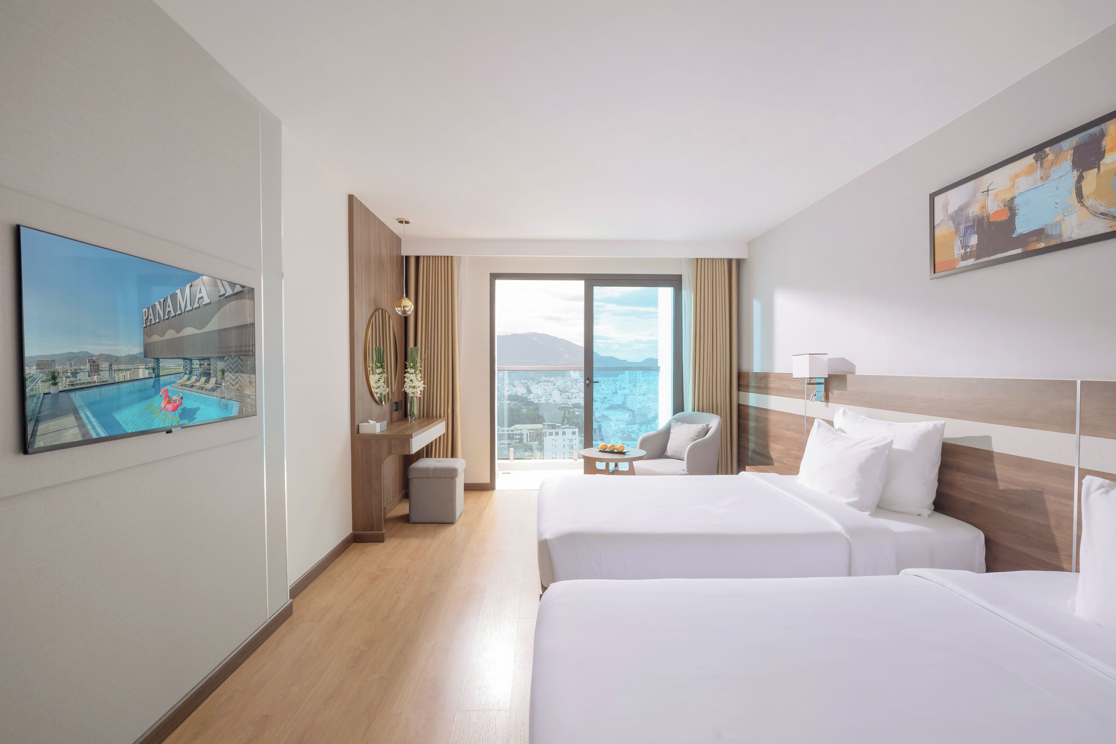 A hotel room with two beds, a desk, a chair, a view of the city, and a painting on the wall.