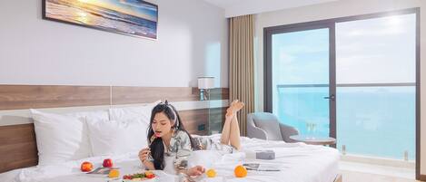 Habitación de hotel con una cama grande, un cuadro de un atardecer, vista al mar y un balcón con una silla.
