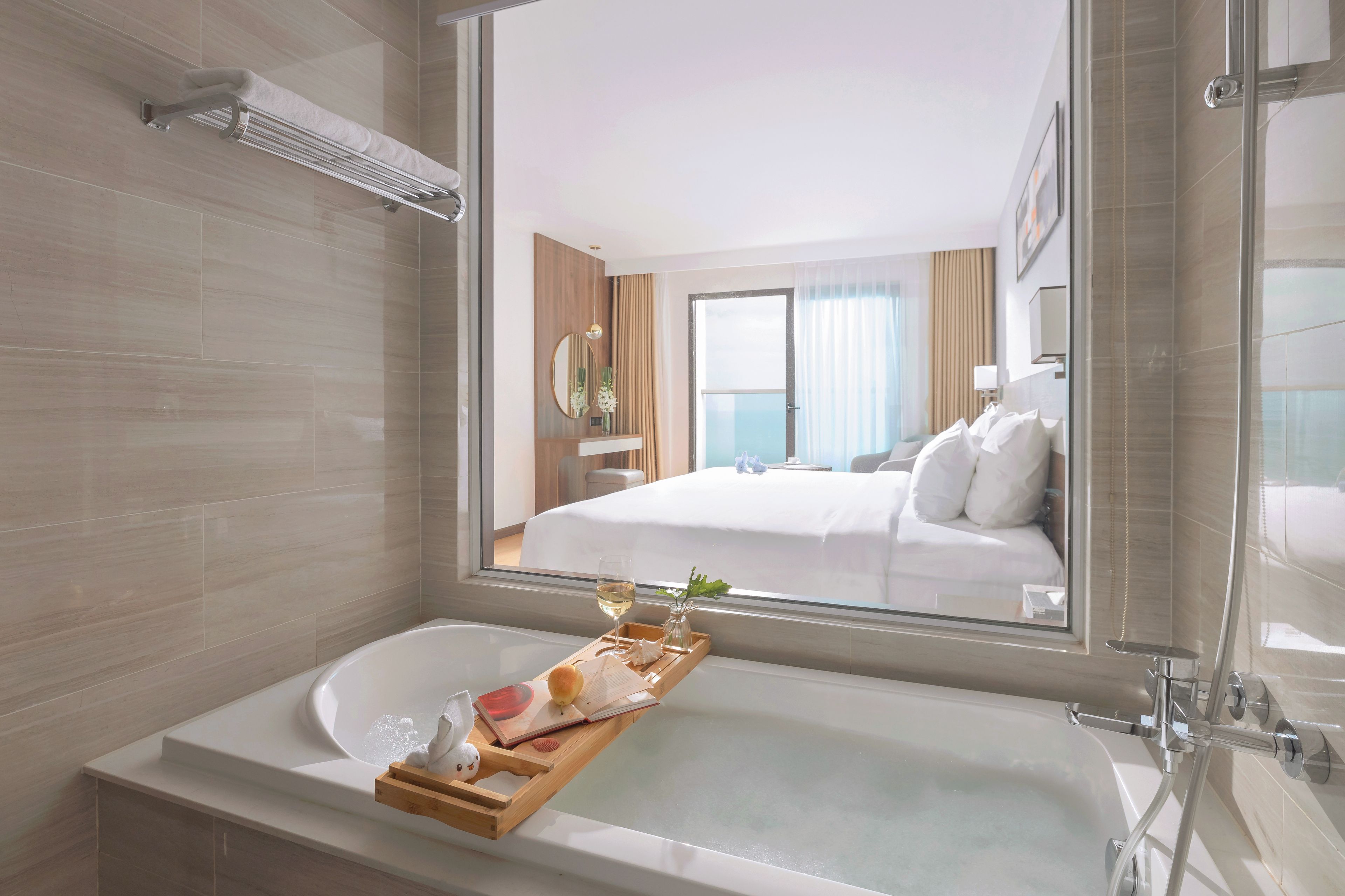 Un bagno moderno con una vasca, un vassoio di legno con un libro e una piccola statuina, e un grande specchio che riflette una camera da letto con un letto e una finestra.