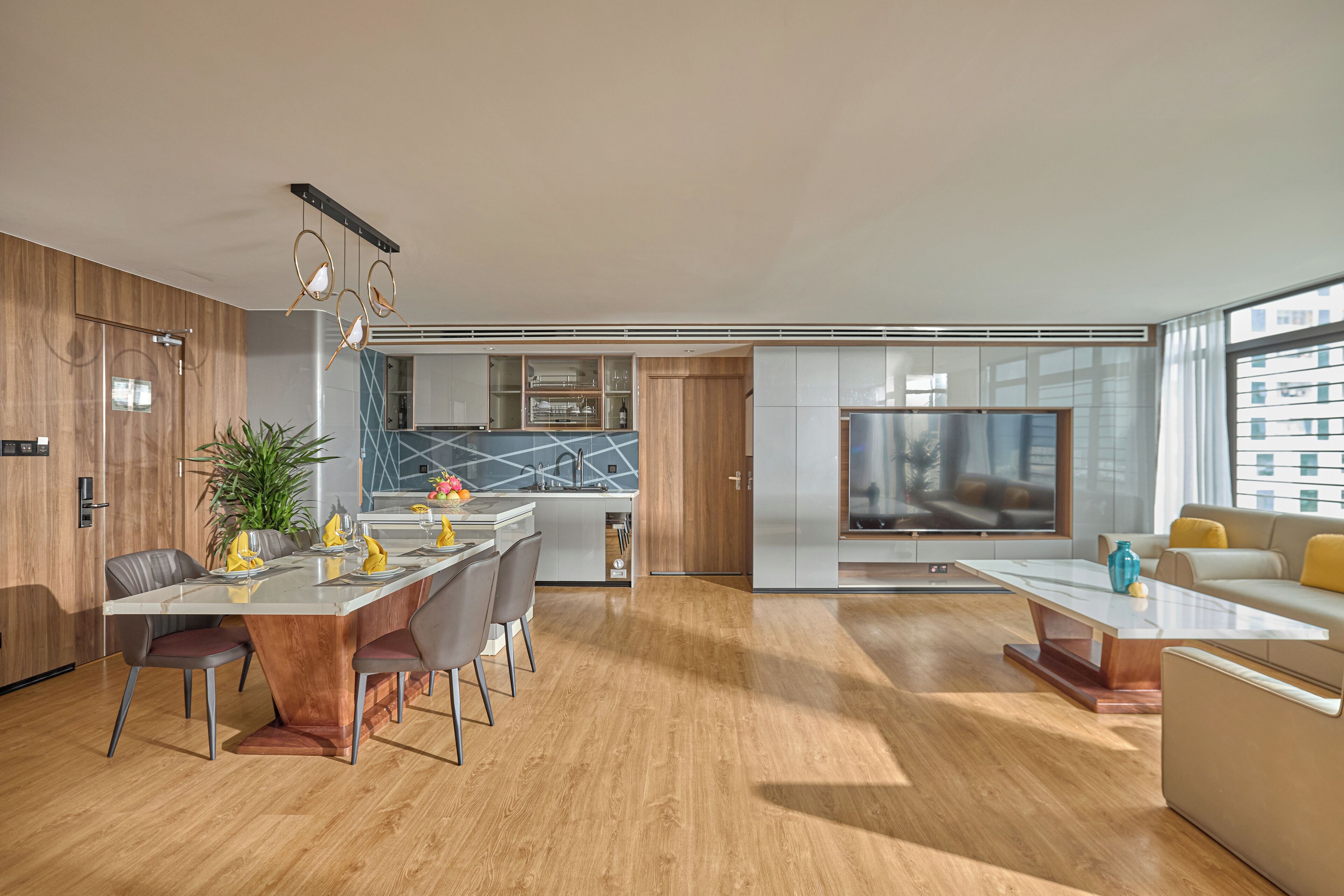 Una sala moderna con un televisor grande, una zona de comedor con una mesa y sillas de madera, y una cocina con horno y microondas integrados.