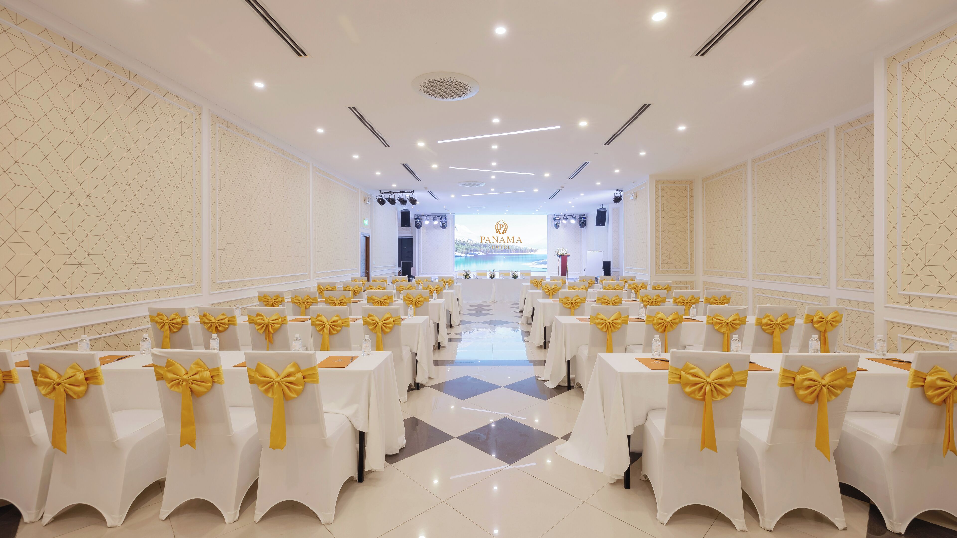 A hotel conference room with rows of white tables and chairs, each with yellow sashes.
