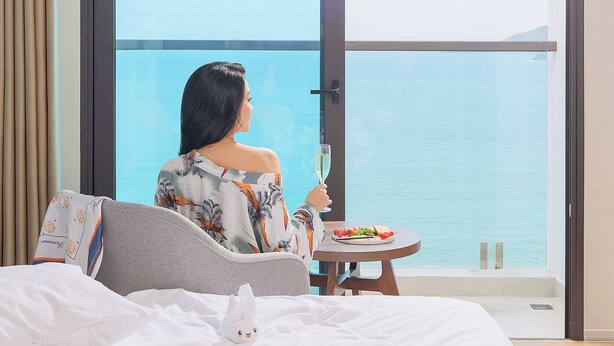 A hotel room with a large window overlooking a body of water, a bed with a magazine, a glass of water, and a small table with a plate of food. - PANAMA Nha Trang Hotel (Nha Trang)