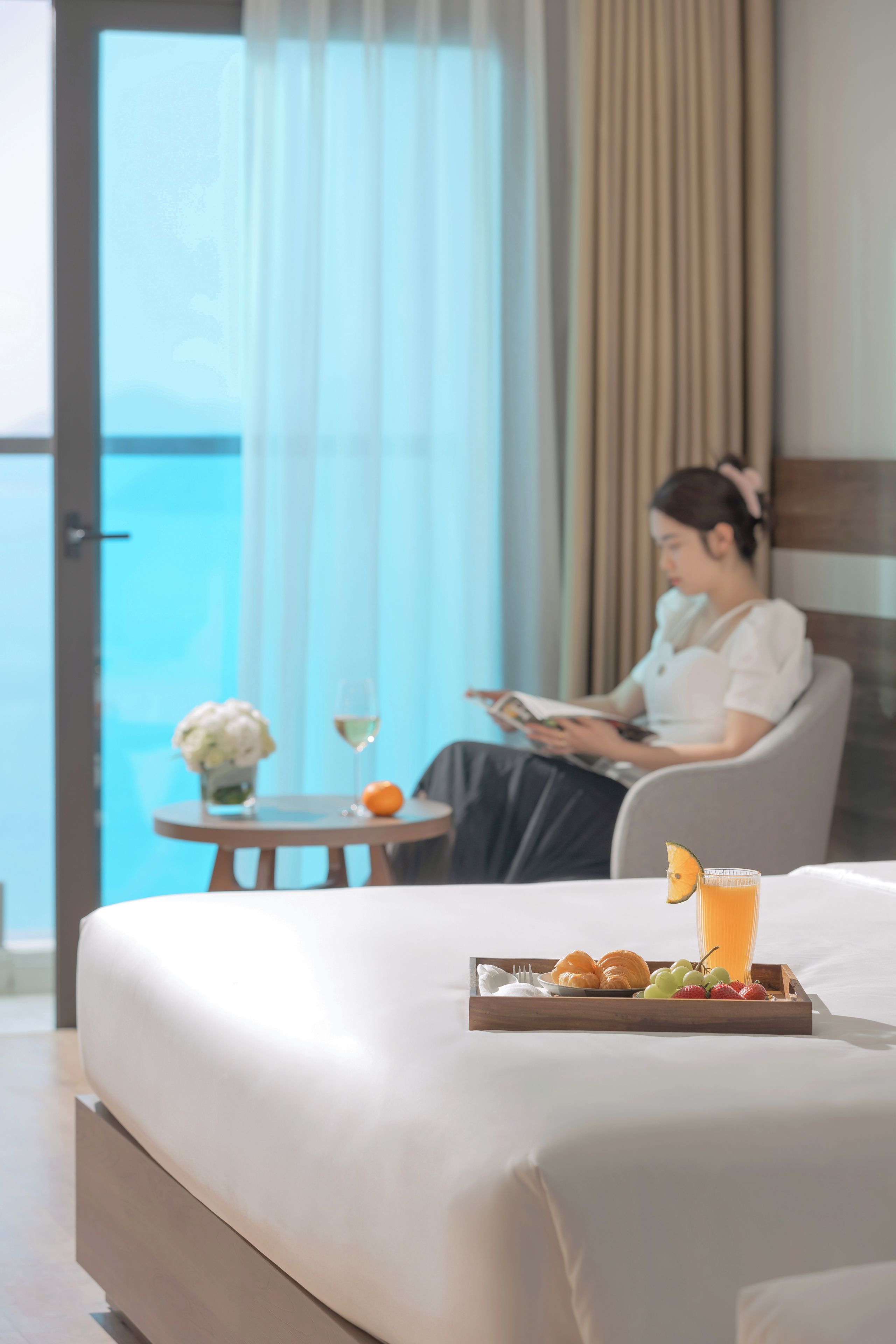 A hotel room with a bed, a dining table with food and drinks, and a view of the sea through a window.
