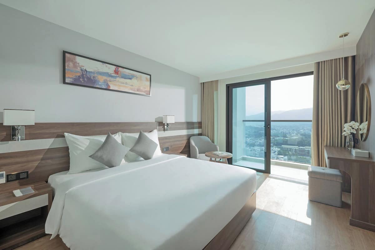 Een moderne hotelkamer met een groot bed, een bureau, een stoel en een uitzicht op de stad door een groot raam.