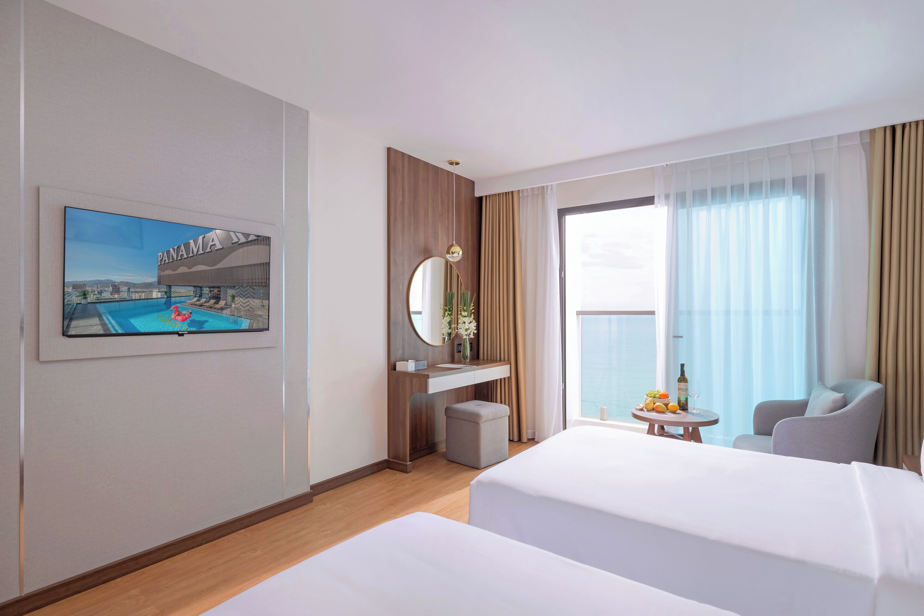 A hotel room with two beds, a large window with curtains, a painting of Panama City, a round mirror, a small table with a bottle and fruit, and a chair.