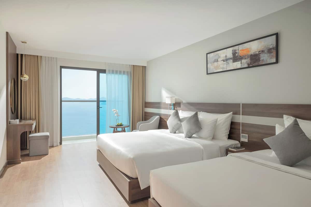 Hotelkamer met twee bedden, een bureau, een stoel, een nachtkastje, een lamp en uitzicht op zee.