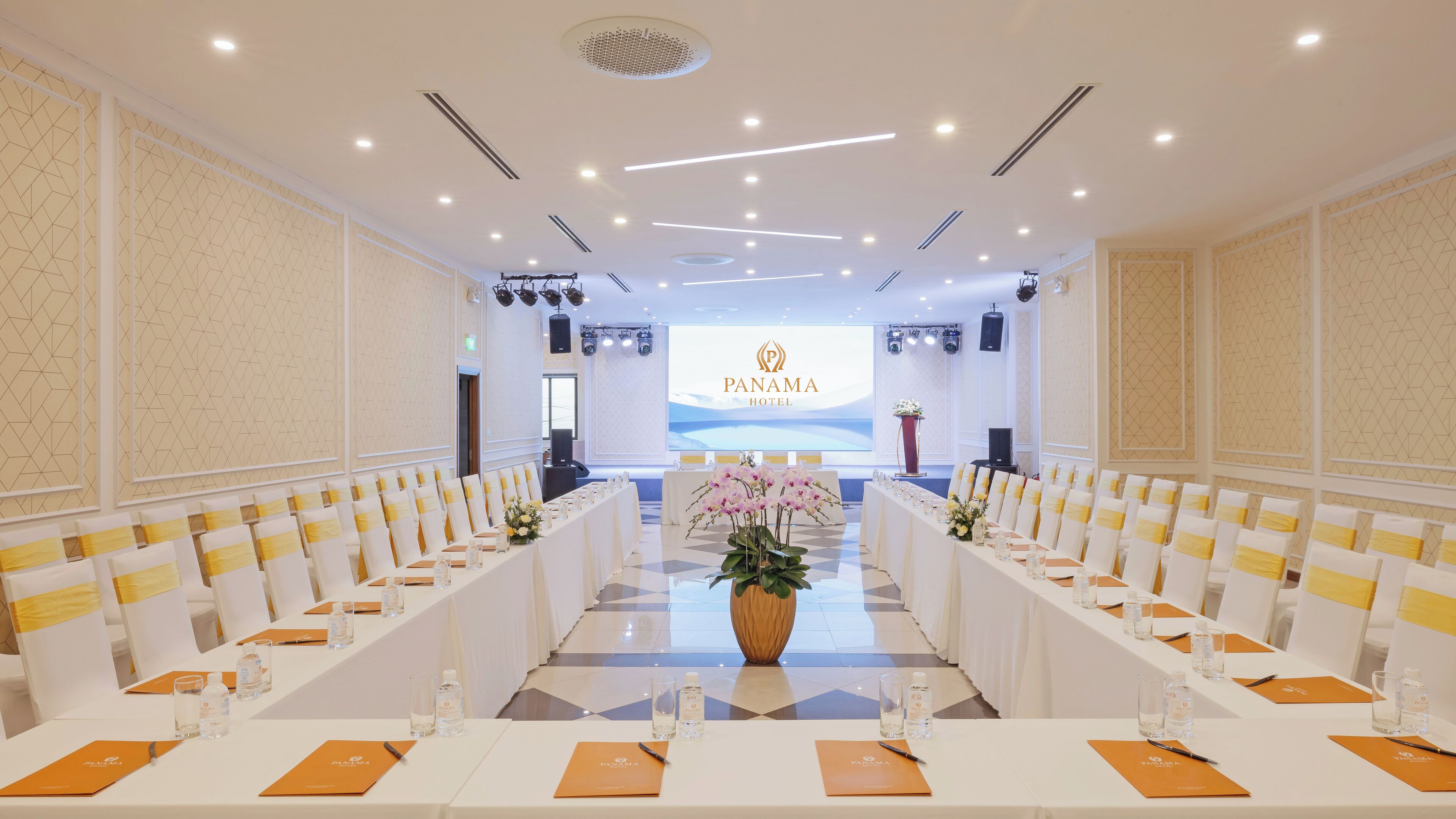 Ein Konferenzraum mit weißen Tischen und Stühlen, einem großen Blumenarrangement und einem Rednerpult mit einem Bildschirm, der das Logo des Hotels zeigt.