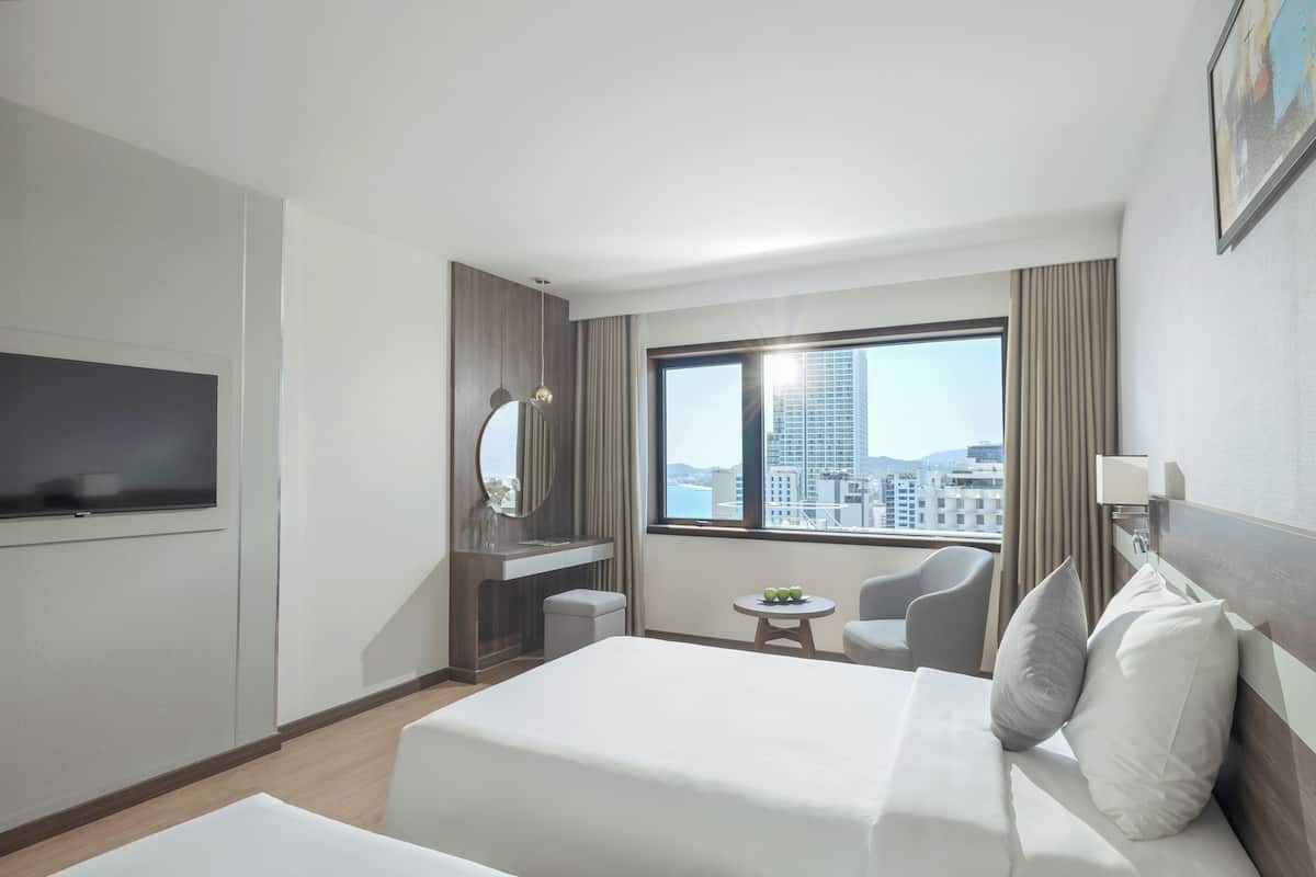 Hotelkamer met een bed, bureau met spiegel, stoel, klein tafeltje met fruit en groot raam met stadszicht.