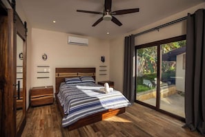 Ein Schlafzimmer mit Bett, Holzboden, Deckenventilator, Schiebeglastür und Blick ins Freie.