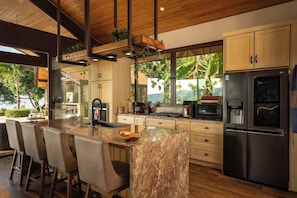 A modern kitchen with a marble island, stainless steel appliances, and a view of greenery outside.