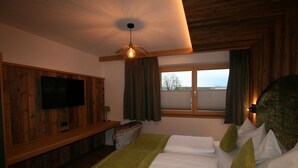A hotel room with a wooden ceiling, a television mounted on the wall, a bed with white linens and green pillows, a bench with a lamp, and a window with curtains. - Cosy apartment for 6 guests with WIFI, TV and terrace (Hart Im Zillertal)