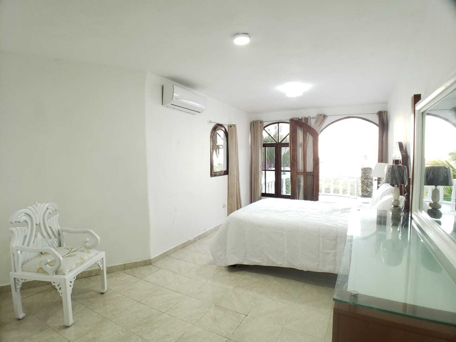 Un dormitorio con cama, una silla blanca, una ventana con cortinas y una mesita de vidrio con lámpara.