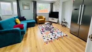 A modern living room with a sofa, a small round table, a TV, and a refrigerator. - 4 Bed House - Sleeps 9 - Garden - Parking - Wifi (Birmingham)