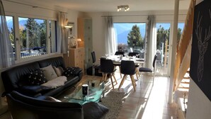 A modern living room with a black leather sofa, a glass coffee table, and a dining area with a table set for four. Large windows offer a view of trees and mountains. - Wonderful  villa for 6 people with WIFI, TV and terrace (Bluche)