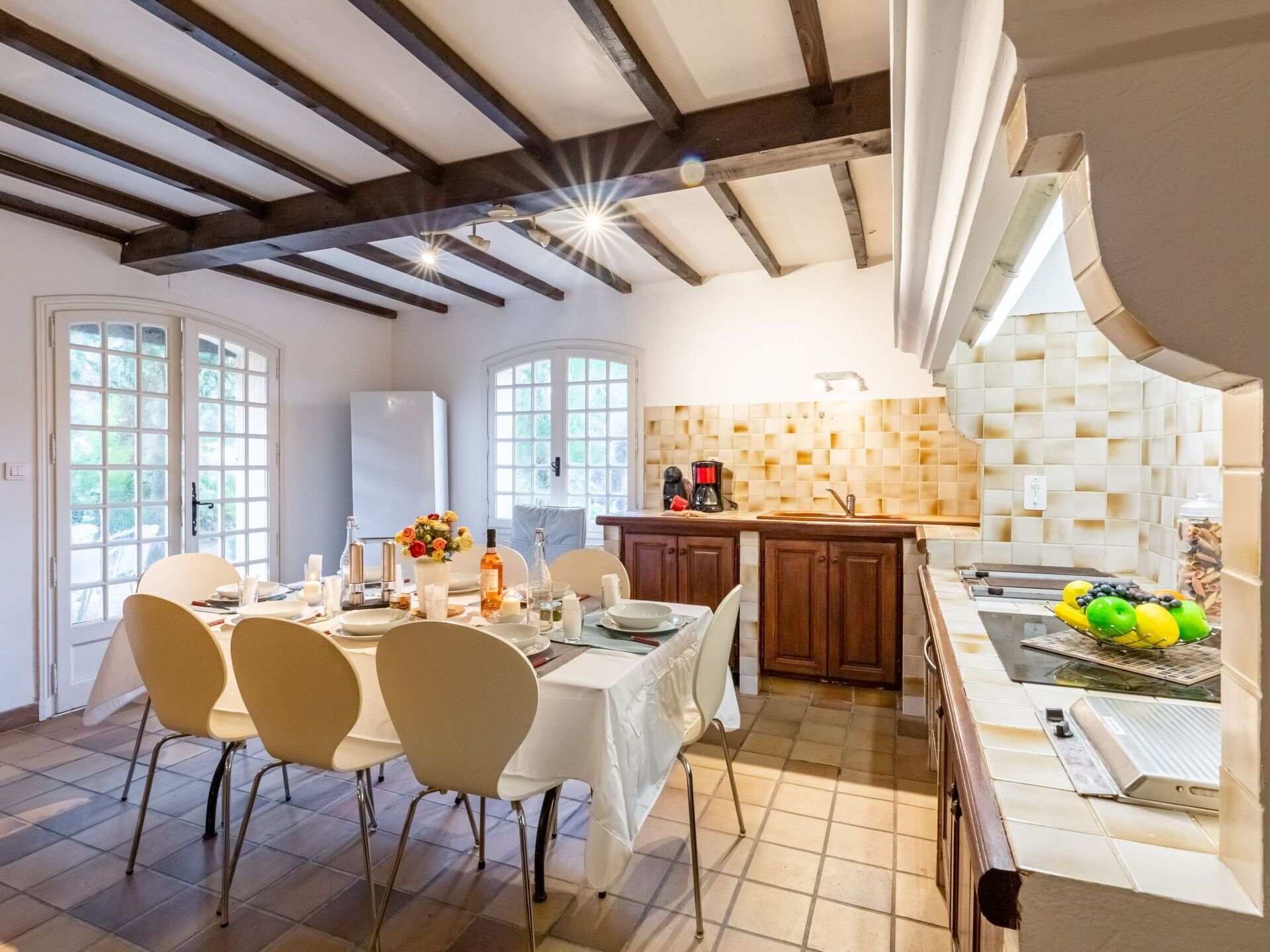 A dining area with a table set for four, a kitchen with a tiled backsplash, and a view of the outdoors through large windows.