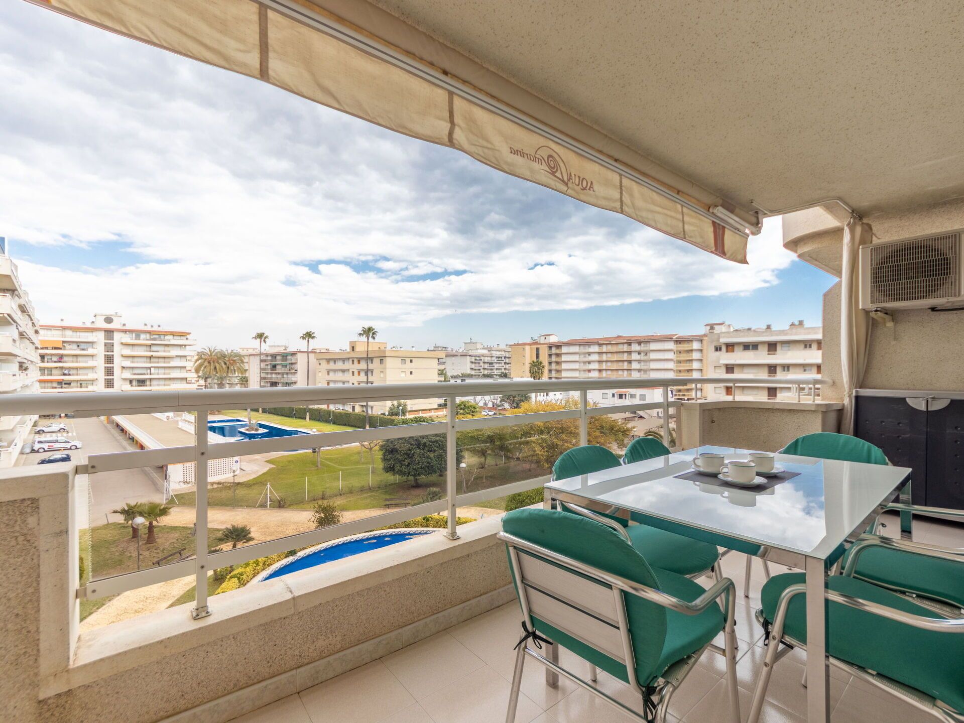 En balkon med et bord og stole, en pool og udsigt til omkringliggende bygninger.