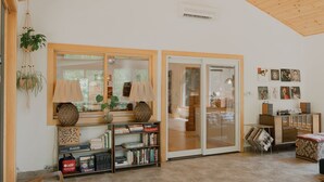 A spacious living room with a wooden ceiling, a large window, a bookshelf, and a sliding glass door leading to another room.