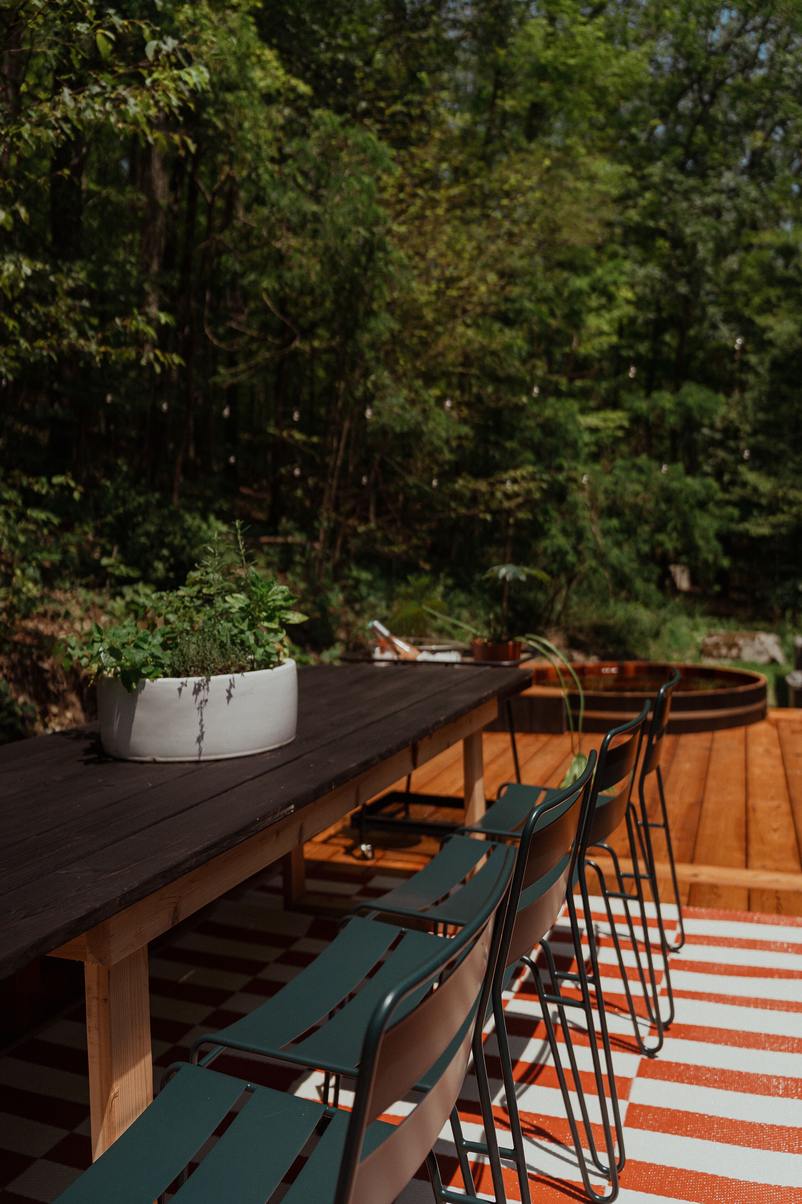 Una terraza de madera con una larga mesa y varias sillas, rodeada de abundante vegetación.