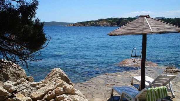 A rocky seaside patio with a wooden umbrella, two lounge chairs, and a stone pathway.