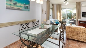 Een moderne kamer met een glazen eettafel, een bed met een geweven hoofdbord, een televisie en een schilderij van een strandgezicht.