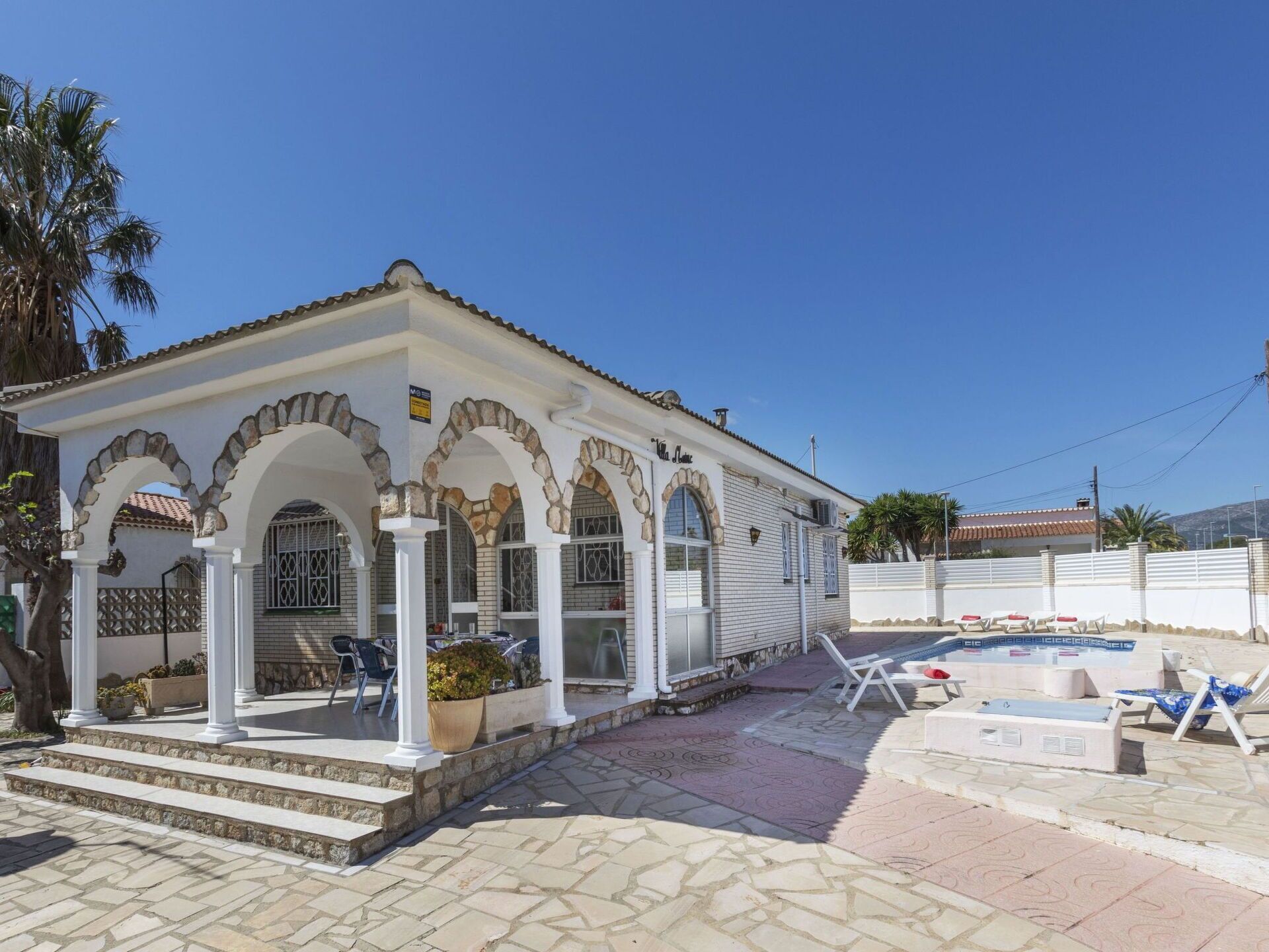 A white building with stone arches and a tiled roof, a pool area with lounge chairs, and a clear blue sky.