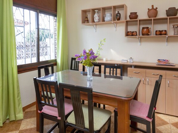 Un comedor con mesa de vidrio, sillas de madera con asientos acolchados, un florero con flores y estanterías con cerámica y utensilios de cocina.