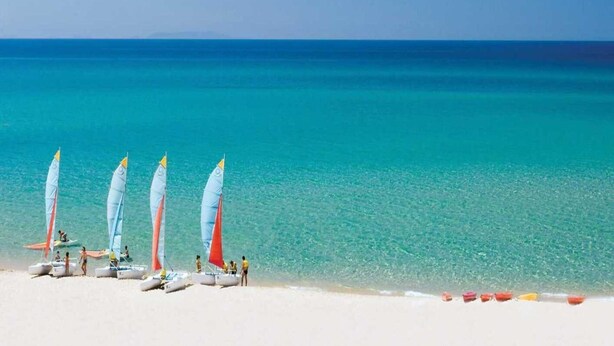 Una playa con varios veleros alineados en la arena, un cielo azul despejado y aguas turquesas tranquilas.