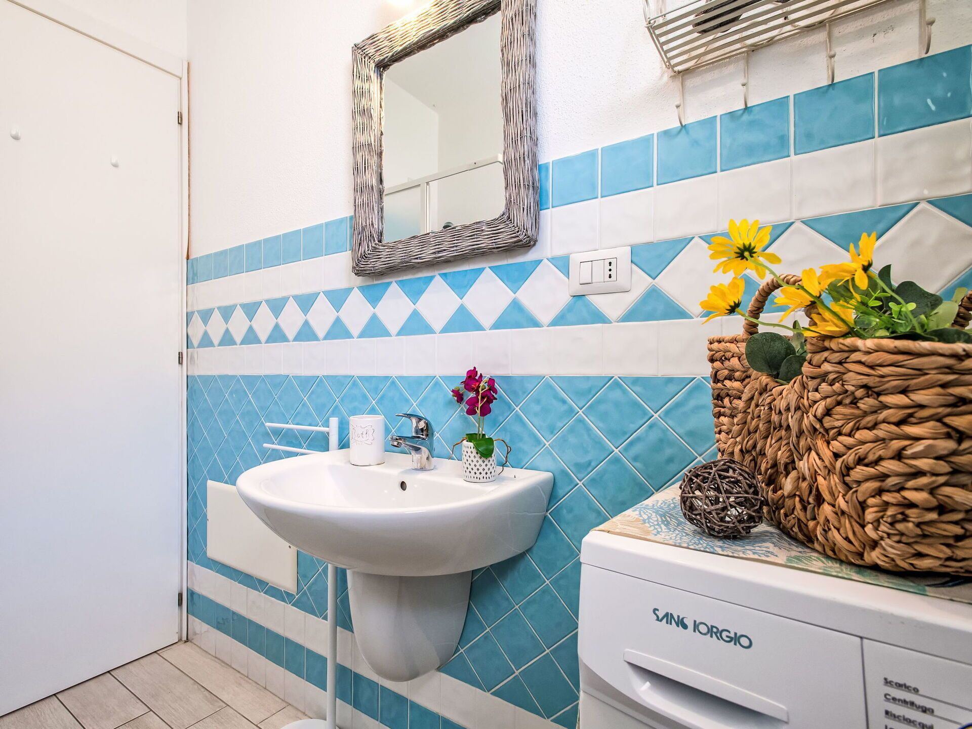 A bathroom with a white sink, blue and white patterned tiles, a mirror, and a basket with flowers.