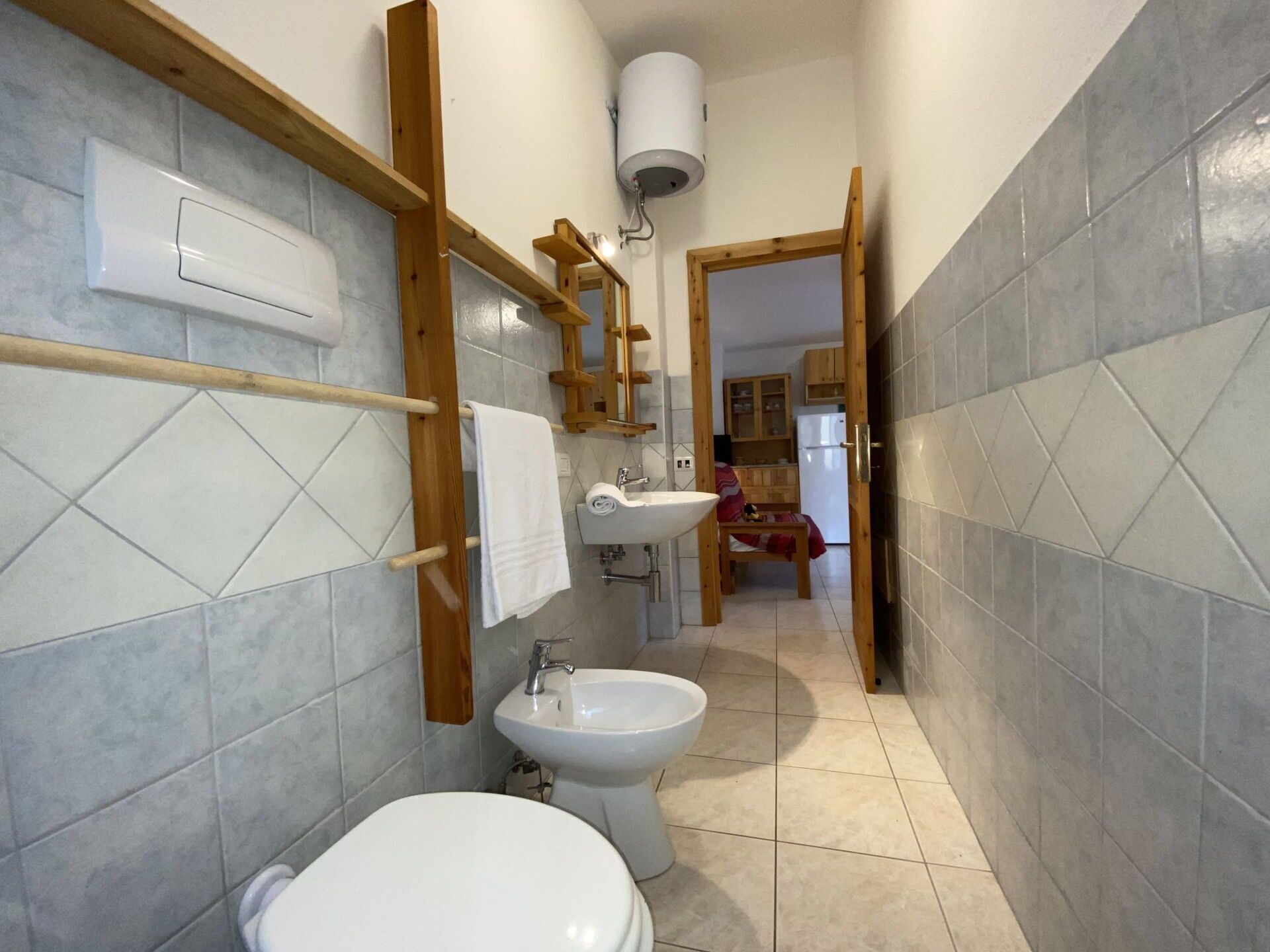 A narrow bathroom with a toilet, bidet, and sink. A towel hangs on a rack, and a wooden shelf is above the sink.
