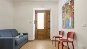 Une pièce avec un canapé bleu, deux fauteuils rouges, une porte en bois et un tableau coloré accroché au mur.