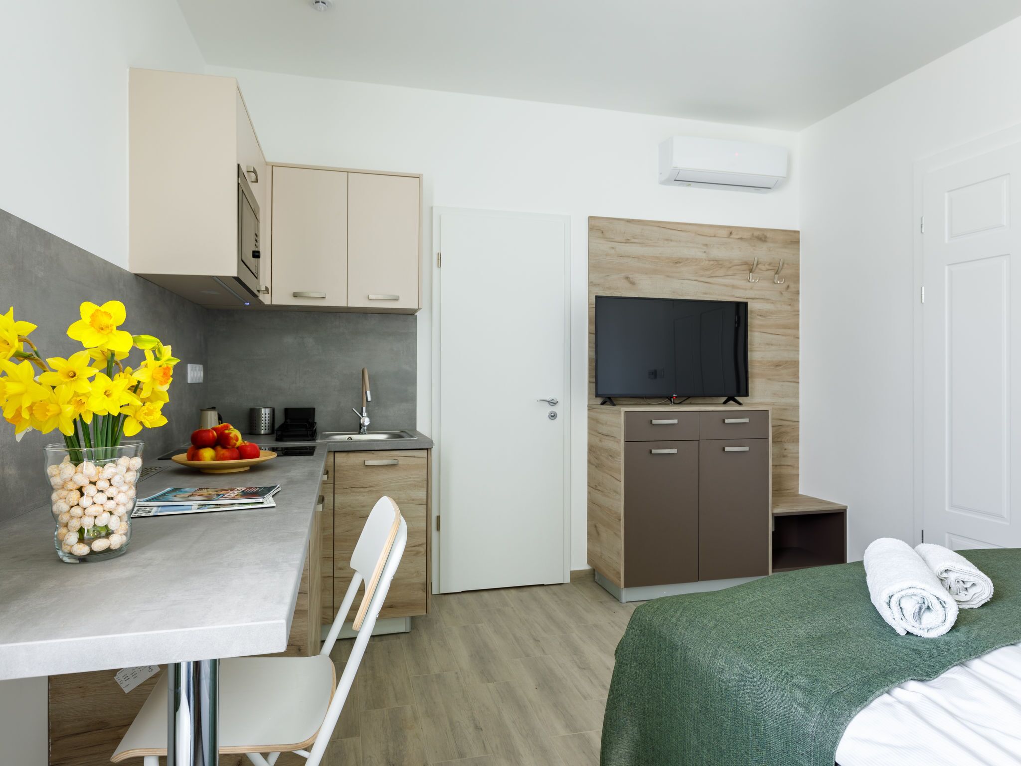 A compact room with a kitchenette, a bed with a green blanket, a TV on a wooden wall, and a vase with yellow flowers.