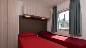 A room with two single beds, a wardrobe, a window with curtains, and a headboard with built-in lighting.