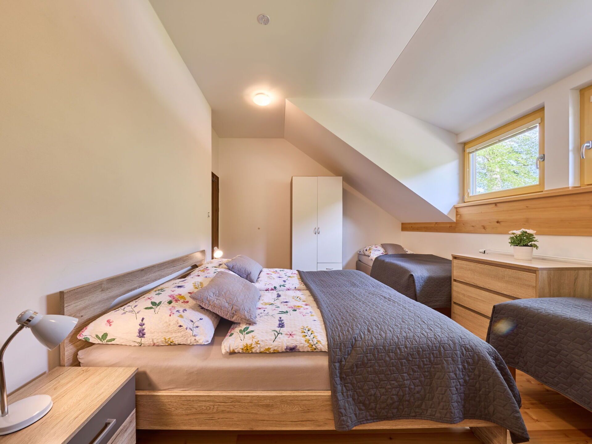 Een slaapkamer met een bed, nachtkastje, lamp en raam met uitzicht op groen.
