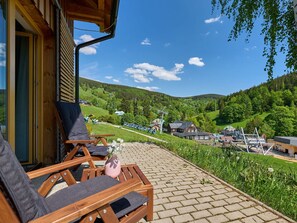 Una terraza de madera con mobiliario exterior y vistas a un valle verde con edificios y un telesilla.