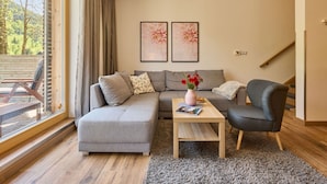 A modern living room with a grey sectional sofa, a wooden coffee table, and a grey armchair. There are two framed floral artworks on the wall, a window with curtains, and a view of trees outside. - Lovely apartment for 4 people with WIFI, TV, terrace, pets allowed and panoramic view (Dvur Králové Nad Labem)