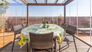 A covered patio with a table and chairs, large windows, and a view of the surrounding area.