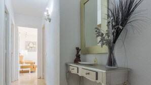 A hallway with a white wall, a light-colored floor, a white console table with a mirror, a vase with dried plants, and a small statue.