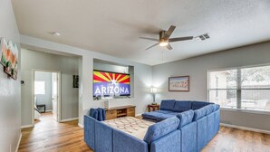 A spacious living room with a large blue sectional sofa, a ceiling fan, and a wall-mounted TV displaying the Arizona state flag. - Mesa Vacation Home - Sleeps 12 - Close to Golf Courses and SO MUCH MORE! (Mesa)