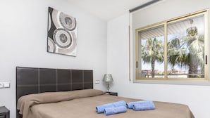 Une chambre à coucher comprenant un lit, une table de chevet, une lampe, une fenêtre donnant sur des palmiers et une œuvre d’art murale.