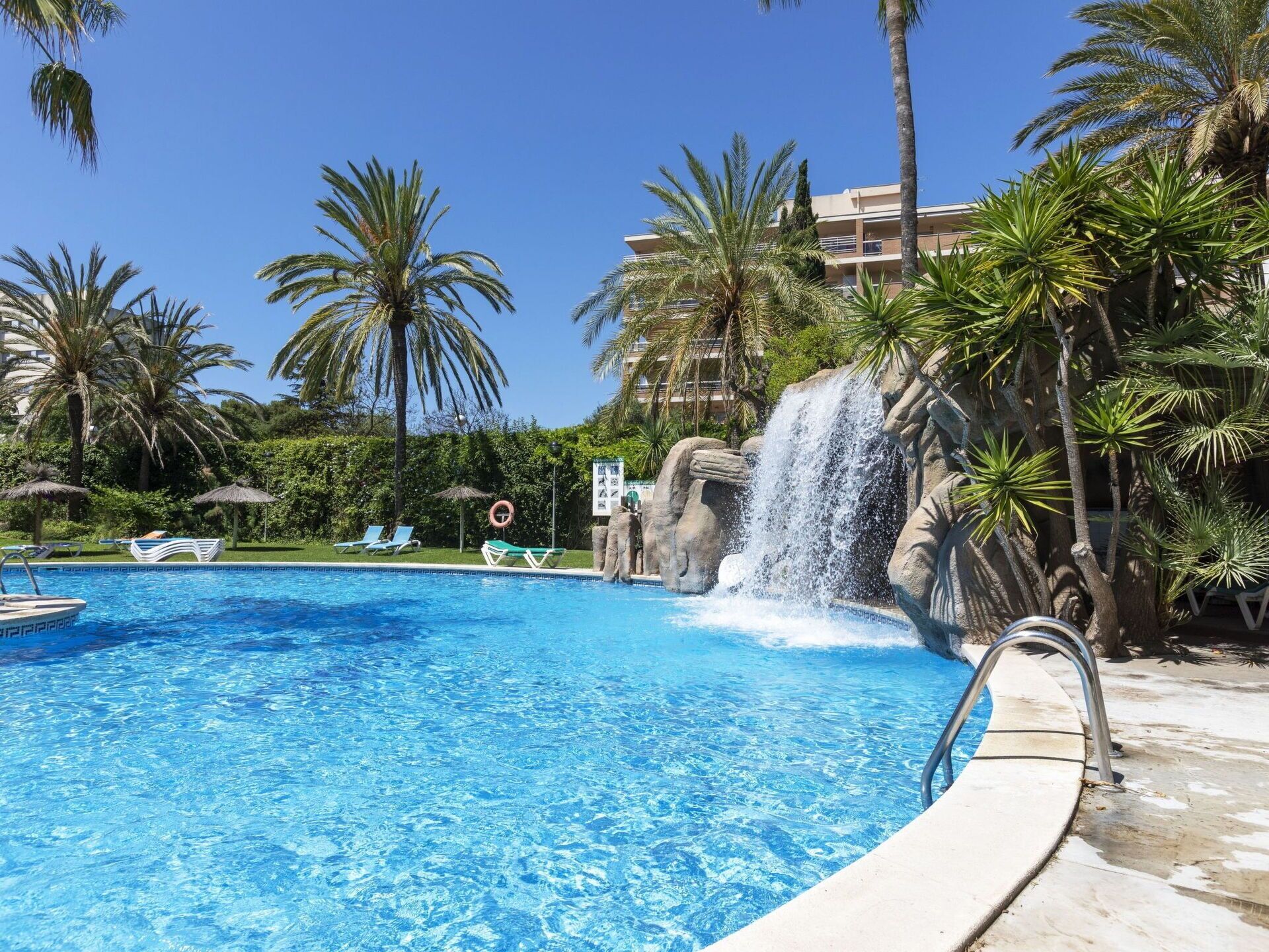 A swimming pool with a waterfall feature, surrounded by palm trees and lounge chairs.