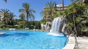 A swimming pool with a waterfall feature, surrounded by palm trees and lounge chairs. - Cosy apartment for 6 guests with A/C, pool, WIFI, terrace and pets allowed (Salou)