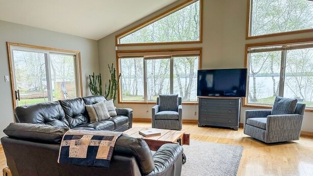 Amplia sala de estar con un sofá de cuero, dos sillones, un televisor de pantalla plana y ventanas amplias que ofrecen vistas a árboles y un lago.