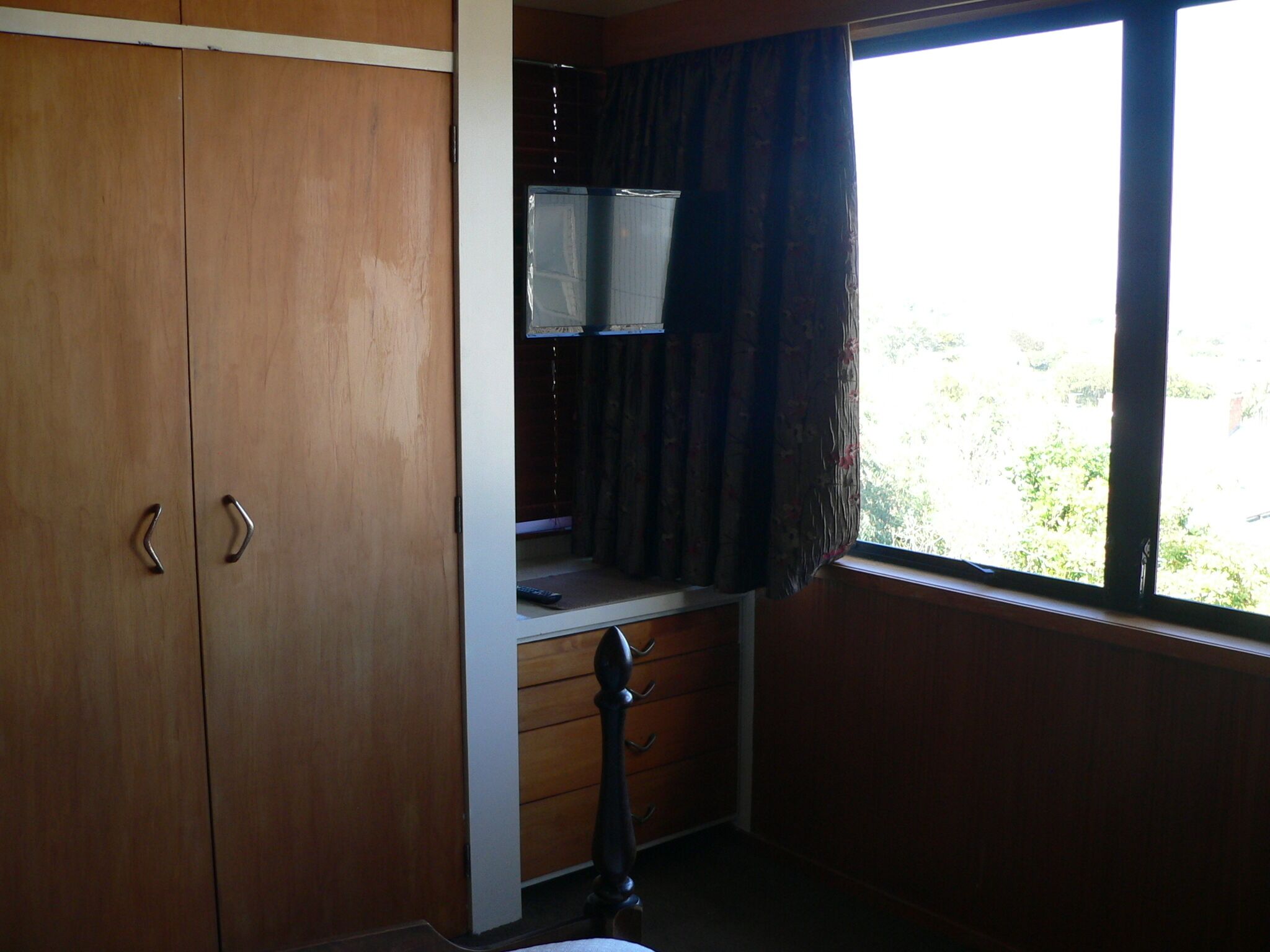A room with a wooden wardrobe, a desk with a chair, and a window with curtains.