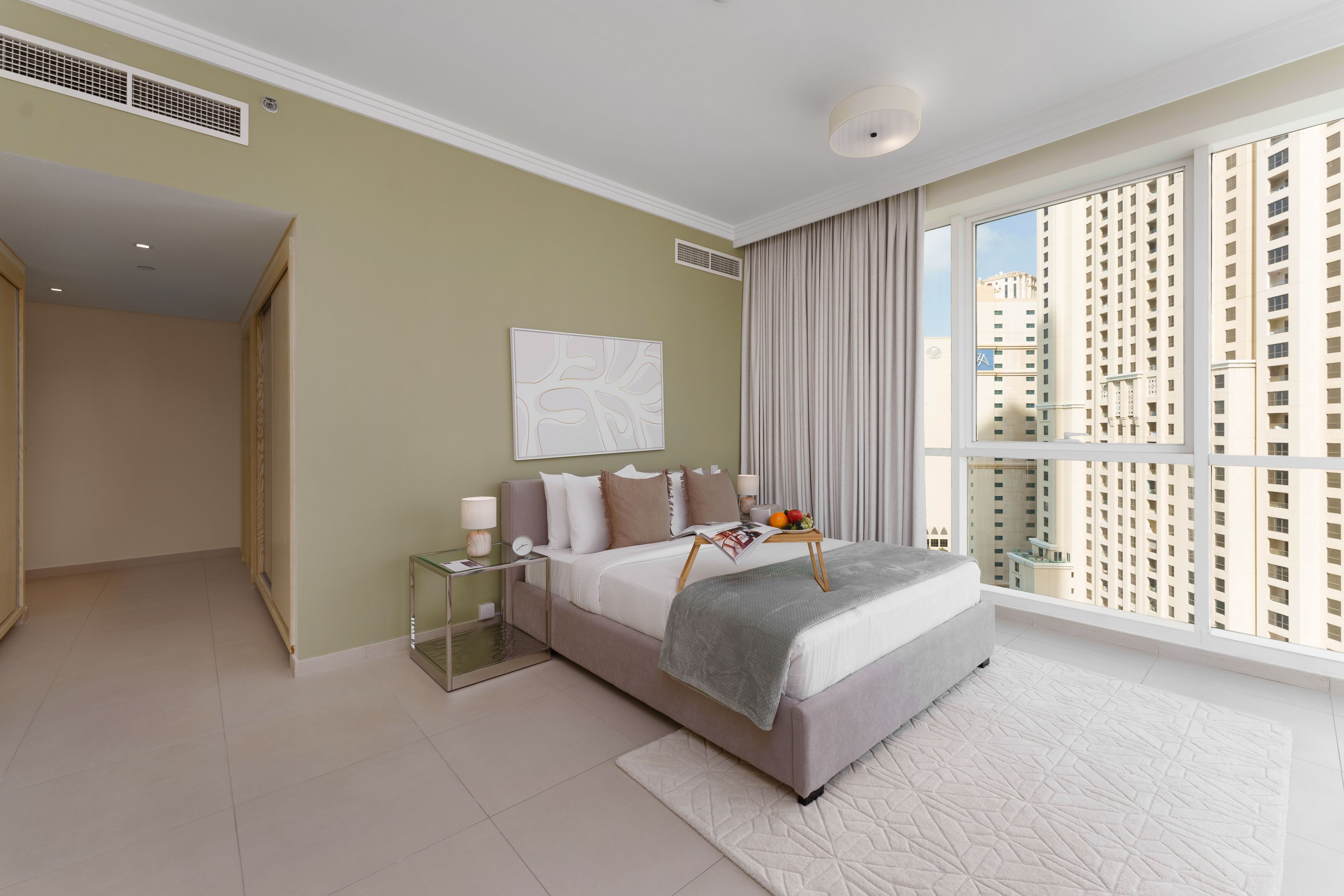 A modern hotel room with a large bed, a bedside table with a lamp, a view of tall buildings through large windows, and a neutral color scheme.