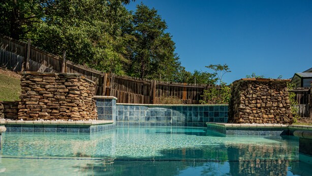 Une piscine aux eaux cristallines, agrémentée de structures en pierre, d’une clôture en bois et d’arbres en arrière-plan.