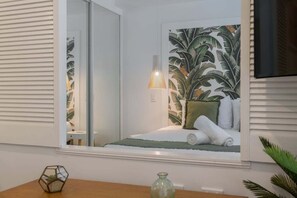 A bedroom with a large window, a bed with white linens, a green throw, and a tropical leaf mural.