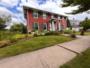 A red brick house with a front porch, a well-maintained lawn, and a pathway leading to the entrance. - The Gibson Grand (North East)
