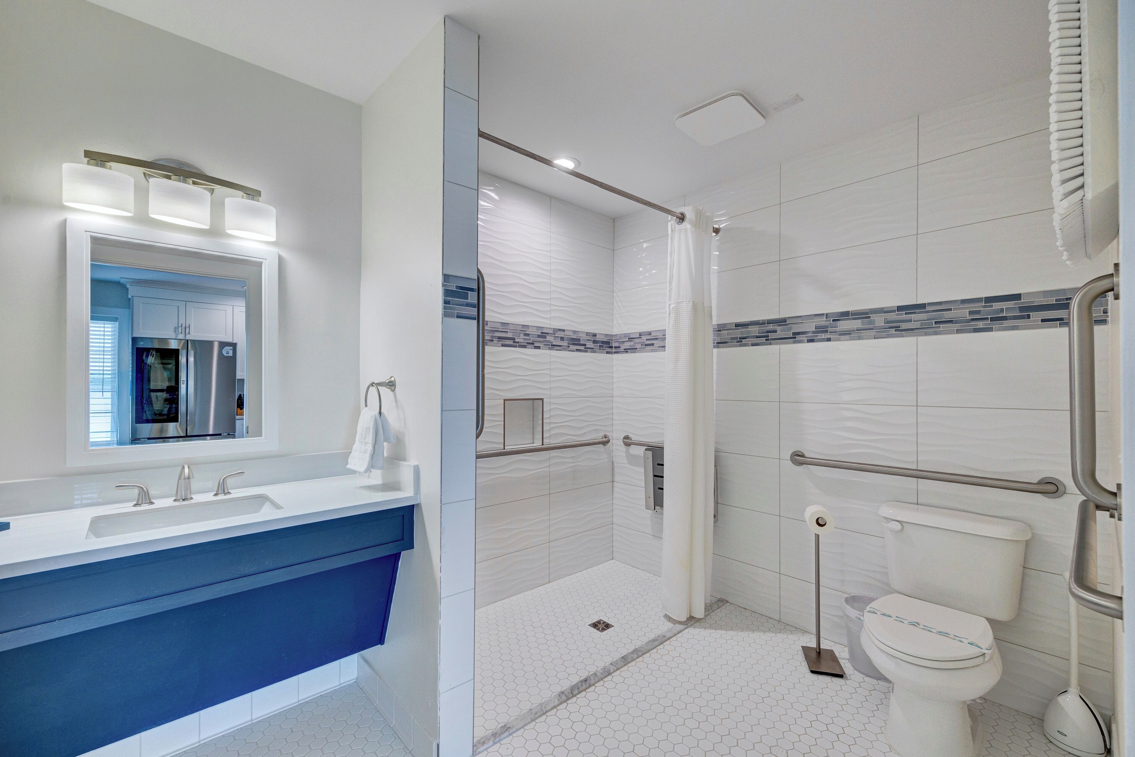 A modern bathroom with a white and blue color scheme, including a double sink, a shower with a glass door, and a toilet with a grab bar.