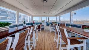 Un área de comedor al aire libre techada, con mesas de madera y sillas blancas, y grandes ventanales que ofrecen vistas a la playa y edificios.